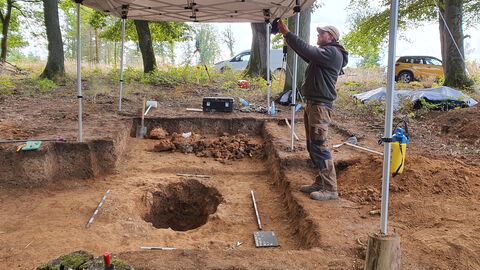 Foto. Ofendokumentation durch einen Mitarbeiter der hessenARCHÄOLOGIE. Im Hintergrund sind erhaltene Fragmente der ehemaligen Ofenröhre zu sehen.