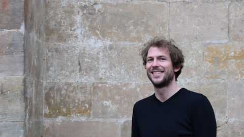 Aufnahme des neuen Bezirksdenkmalpfleger Dr. Malte Nettekoven lächelnd vor einer Wand aus Natursteinen. Sie ist Teil der Elisabethenkirche in Marburg. 