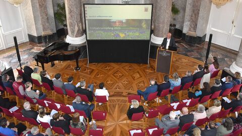 Blick von oben auf eine Zuschauermenge. Oben mittig im Bild befindet sich eine Leinwand, auf der eine Präsentation abgespielt wird. Rechts neben der Leinwand steht Prof. Dr. Markus Harzenetter an einem Rednerpult.