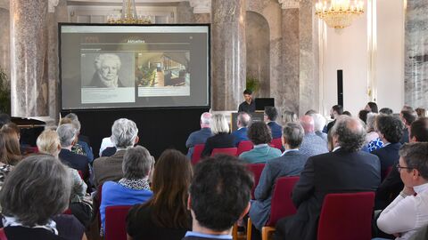 Blick durch das Publikum auf eine Leinwand, auf der eine Präsentation gezeigt wird. Rechts neben der Leinwand steht ein Mann am Rednerpult.