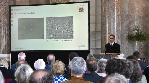 Blick durch das Publikum auf eine Leinwand, auf der eine Präsentation gezeigt wird. Rechts neben der Leinwand steht ein Mann am Rednerpult.