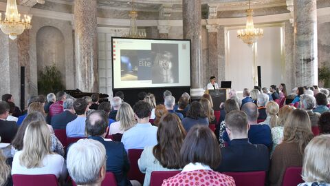 Blick durch das Publikum auf eine Leinwand, auf der eine Präsentation gezeigt wird. Rechts neben der Leinwand steht eine Frau am Rednerpult.
