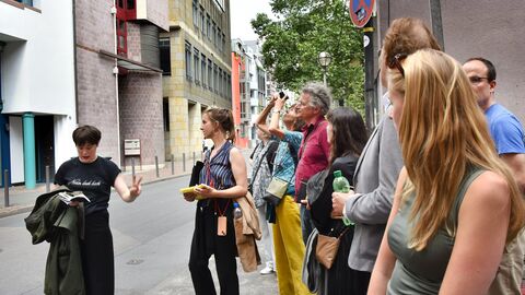 Frau (Helene Peters) steht vor einer Grupppe und erzählt etwas.