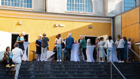 Zur feierlichen Eröffnung des Theaters kamen viele Interessierte. Das neue Kulturzentrum wird gut von der Gemeinde angenommen. Menschen stehen an Tischen vor dem Gebäude und feiern die Eröffnung