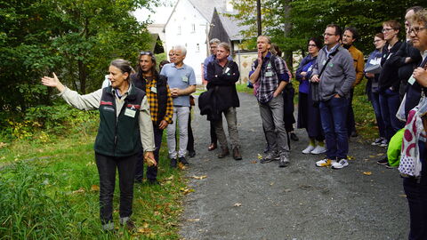 Nach dem Mittagessen wurde das Tagungsthema auf einem Rundgang durch den Hessenpark vertieft. Gruppe steht in einem kleinen Wald, eine Frau spricht zu der Gruppe.
