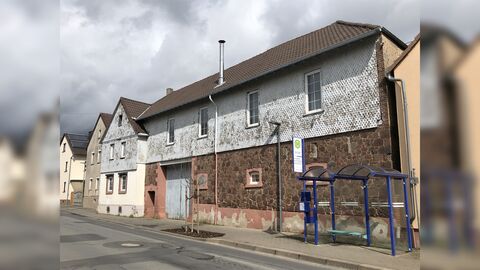 Links zweigeschossiges giebelständiges Wohnhaus mit hellverputztem Erdgeschoss und Holzschindeln verkleidetem Obergeschoss und Giebel. Rechts anschließend langgestreckter traufständiger Längsbau mit Tuffsteinmauerwerk mit Holzschindeln verkleidetem Obergeschoss. 