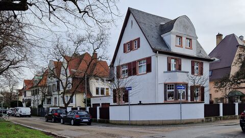 Man sieht ein hellblau verputztes Eckhaus. Es weist die typischen Elemente (hoher Giebel, Erker, Gauben und Fensterläden) der Villenkolonie Dornbusch auf.