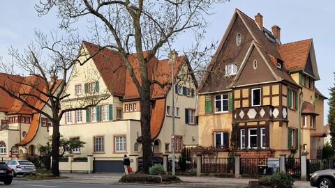 Man sieht den Ausschnitt eines Straßenzuges der Gartenstadt Frankfurt mit drei Villen. Die Gebäude bestechen vor allem  durch ihre hohen Dächer mit den variantenreich ausgeformten Giebeln, Dachgauben, Erkern und Fenstern.  