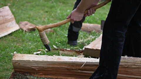 Mit handgefertigten Werkzeugen nach historischem Vorbild werden die gespaltenen Stämme in Form gebracht. Arbeit mit handgefertigtem Werkzeug an einem gespaltenen Stamm.