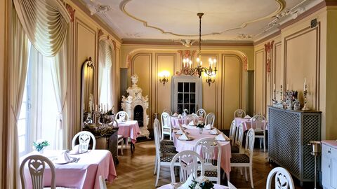 Goßer Raum mit gelb-braunen Wandvertäfelungen. Im Raum stehen mehrere Tische mit pinker Tischdecke und weiße Holzstühle. An der Decke befinden sich Stuckverzierungen. 