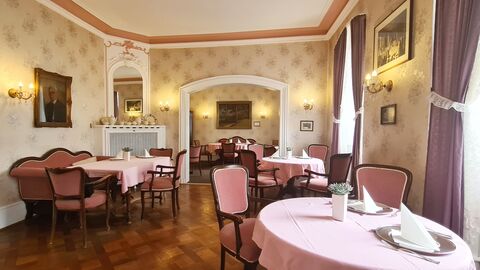 Großer Raum mit gelblicher Stofftapete. Drei Sitzgruppen mit altrosa bezogenen Sesseln und Sofas verteilen sich im Raum. Die Tische haben eine rosafarbene Tischdecke. Durch einen weißen Torbogen blickt man in einen zusätzlichen Raum mit einer weiteren Sitzgruppe.