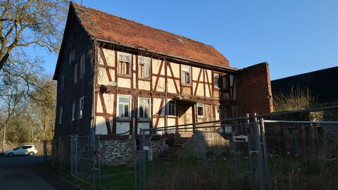 Die Eingangsfassade des Gebäudes mit Rest vom Stallgebäude Fachwerkhaus auf Natursteinsockel mit mittig gelegenem Eingang. An das Gebäude schließt der Rest einer Ziegelmauer an.