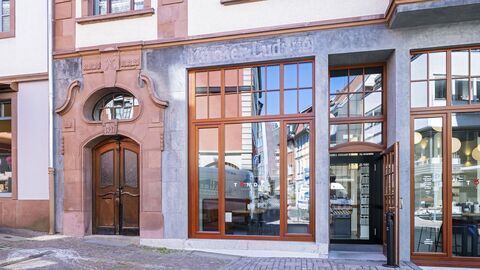 Im Zuge der Fassadensanierung wurden großen Schaufenster wiederhergestellt und die Steinputzfassade freigelegt. Hohe Holzfenster in Steinputzfassade. Ein Schriftzug des ursprünglichen Geschäfts ist oberhalb eines Fensters erkennbar.